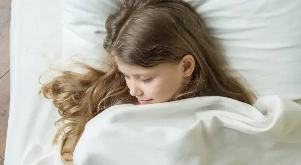 Niña rubia duerme acurrucada, quizás soñando con terrores nocturnos. Su cabello se esparce sobre la almohada blanca.