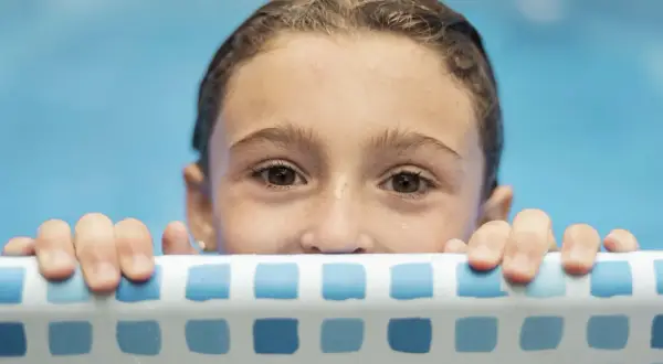 Niña con ojos curiosos se aferra al borde de la piscina, lista para aprender como enseñar a nadar a un niño.