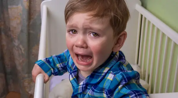 Un niño pequeño con pijama a cuadros azules llora desconsoladamente en su cuna. Mi bebé llora con mucha rabia, con lágrimas corriendo por sus mejillas.