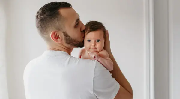 Un padre besa la cabeza de su bebé, que mira a la cámara con sus grandes ojos azules.