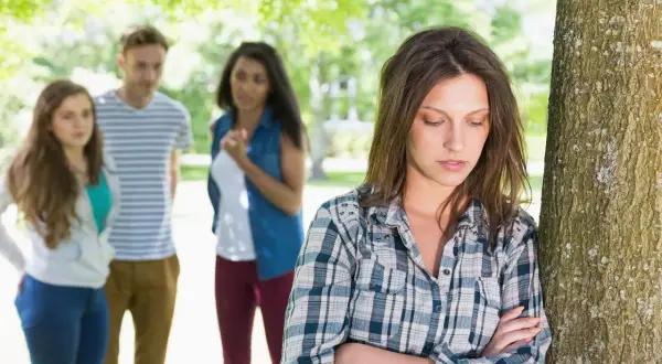 Una chica se siente sola y triste, apoyada en un árbol, mientras sus amigos hablan en el fondo. Es un momento típico de cuando empieza la adolescencia.