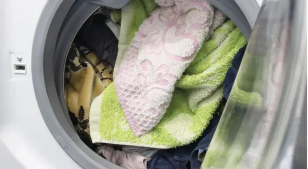Interior de lavadora con ropa variada: toallas verdes y rosas, mantas y prendas oscuras. ¿Cuánto tiempo puede estar la ropa en la lavadora?