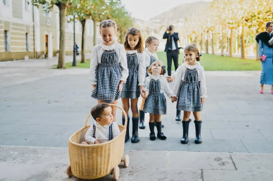 Niños de arras, con vestidos de cuadros y botas, pasean sonrientes. Un bebé en una cesta de mimbre les acompaña.