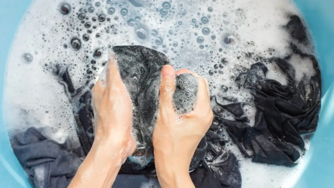 Manos lavando ropa oscura en una tina con espuma. Se puede lavar la ropa con gel de baño para una limpieza suave.