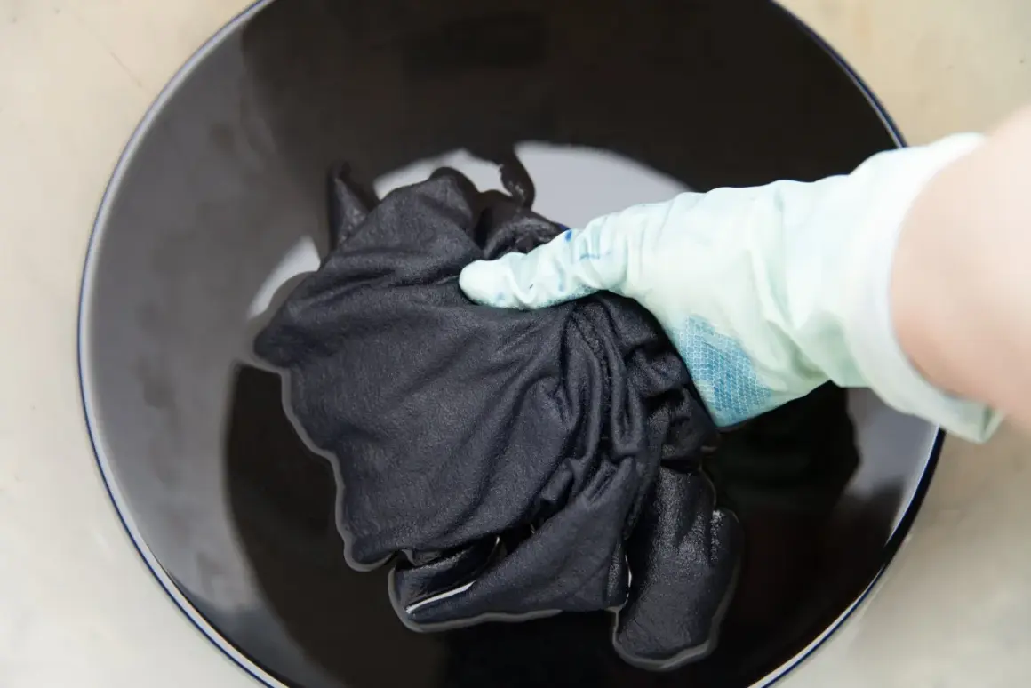 Mano con guante sumerge tela negra en líquido oscuro para recuperar color de ropa oscura.