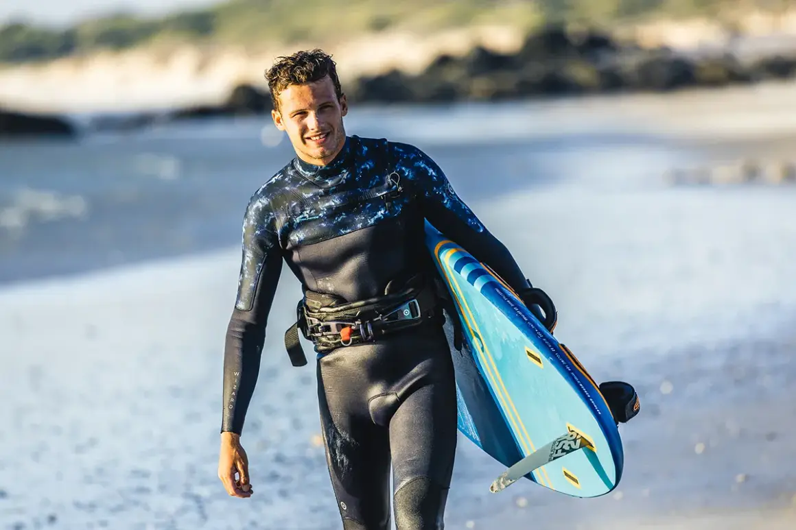 Hombre sonriente con tabla de kitesurf en la playa. Su traje de neopreno es impermeable, ideal para deportes acuáticos.