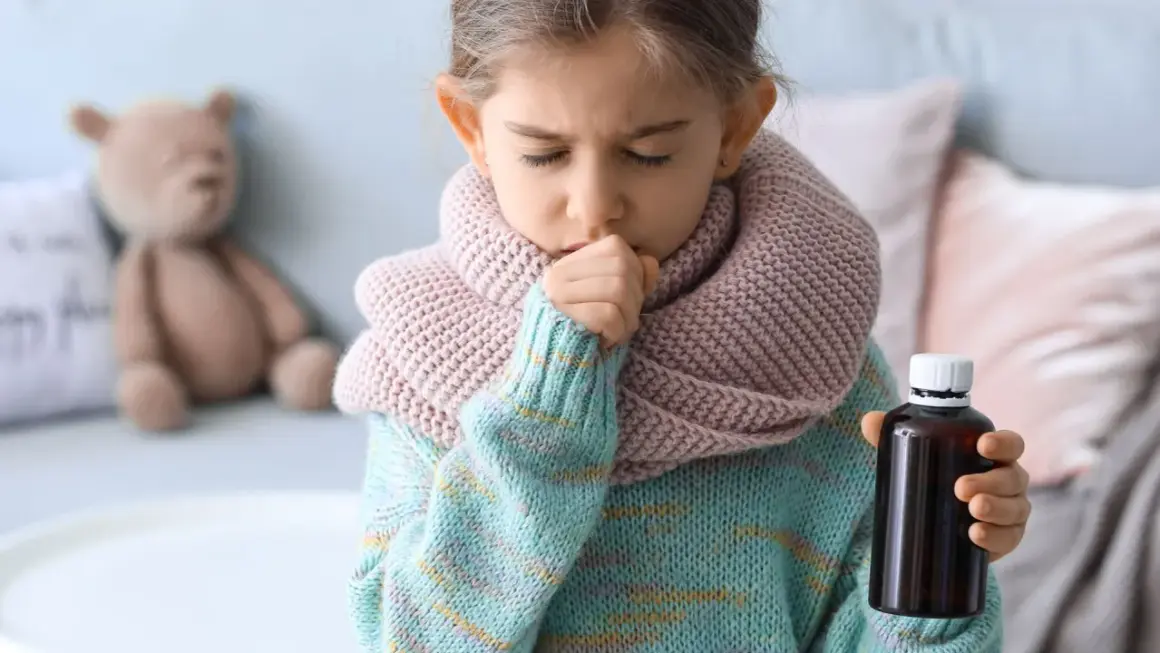 Niña con tos y bufanda, sosteniendo un frasco de medicina. Una imagen que ilustra cómo calmar la tos en niños.
