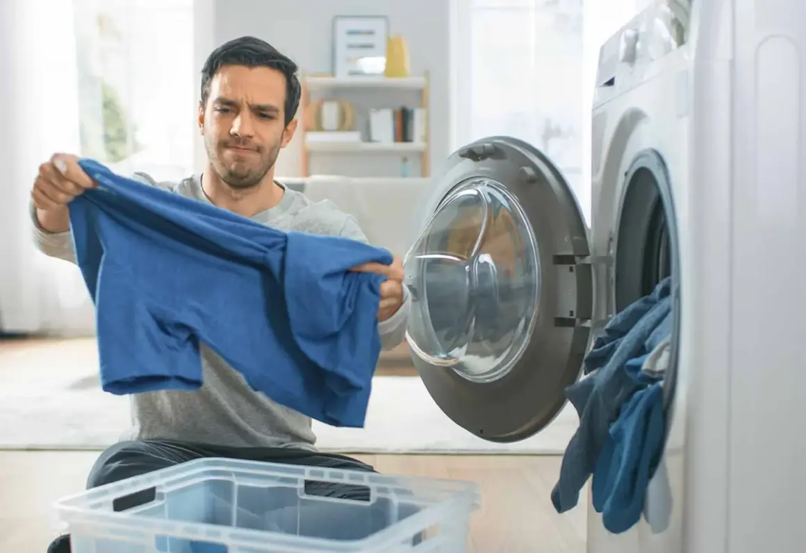 Hombre preocupado revisa una camiseta azul, pensando en como lavar algodón para que no encoja. Cerca, una lavadora abierta con ropa.