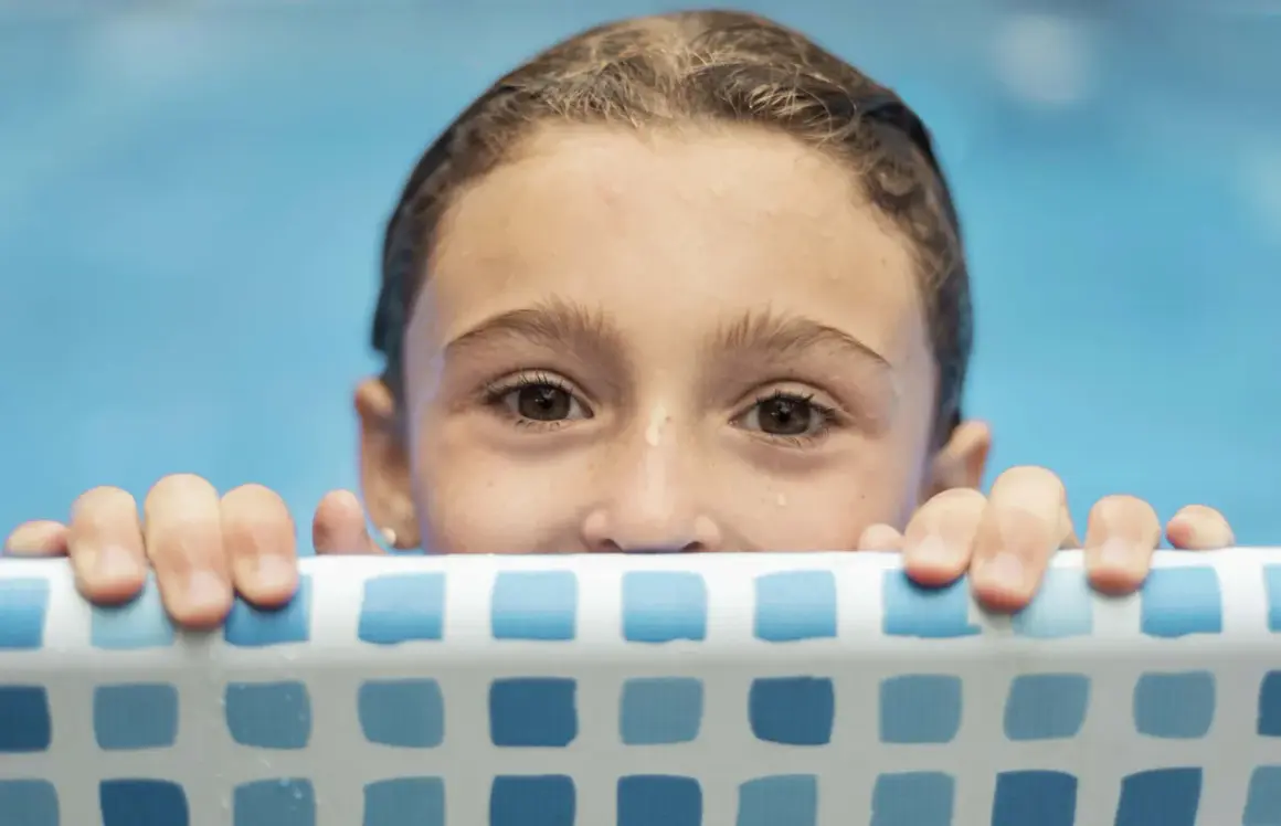 Niña con ojos curiosos se aferra al borde de la piscina, lista para aprender como enseñar a nadar a un niño.