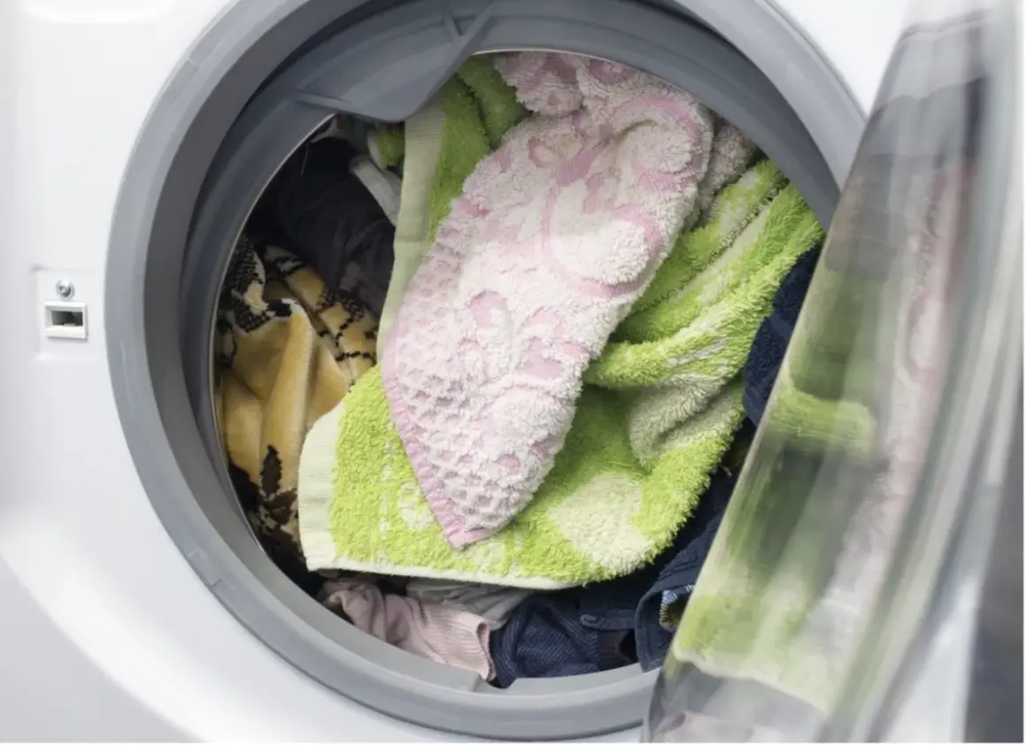 Interior de lavadora con ropa variada: toallas verdes y rosas, mantas y prendas oscuras. ¿Cuánto tiempo puede estar la ropa en la lavadora?