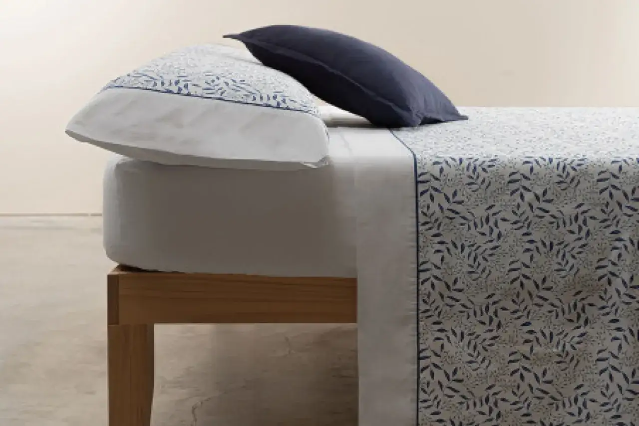 Cama con sábanas de poliéster o microfibra con estampado de hojas azules. Una almohada azul marino y otra con el mismo estampado.