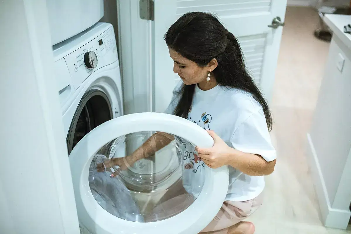 Mujer saca ropa de lavadora, buscando quitar manchas grises.