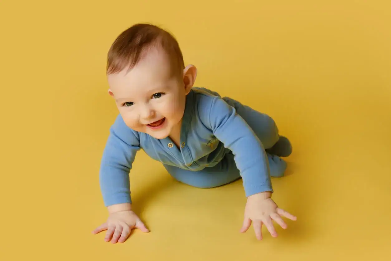 Un beb&eacute; sonriente con un mameluco azul gatea sobre un fondo amarillo.