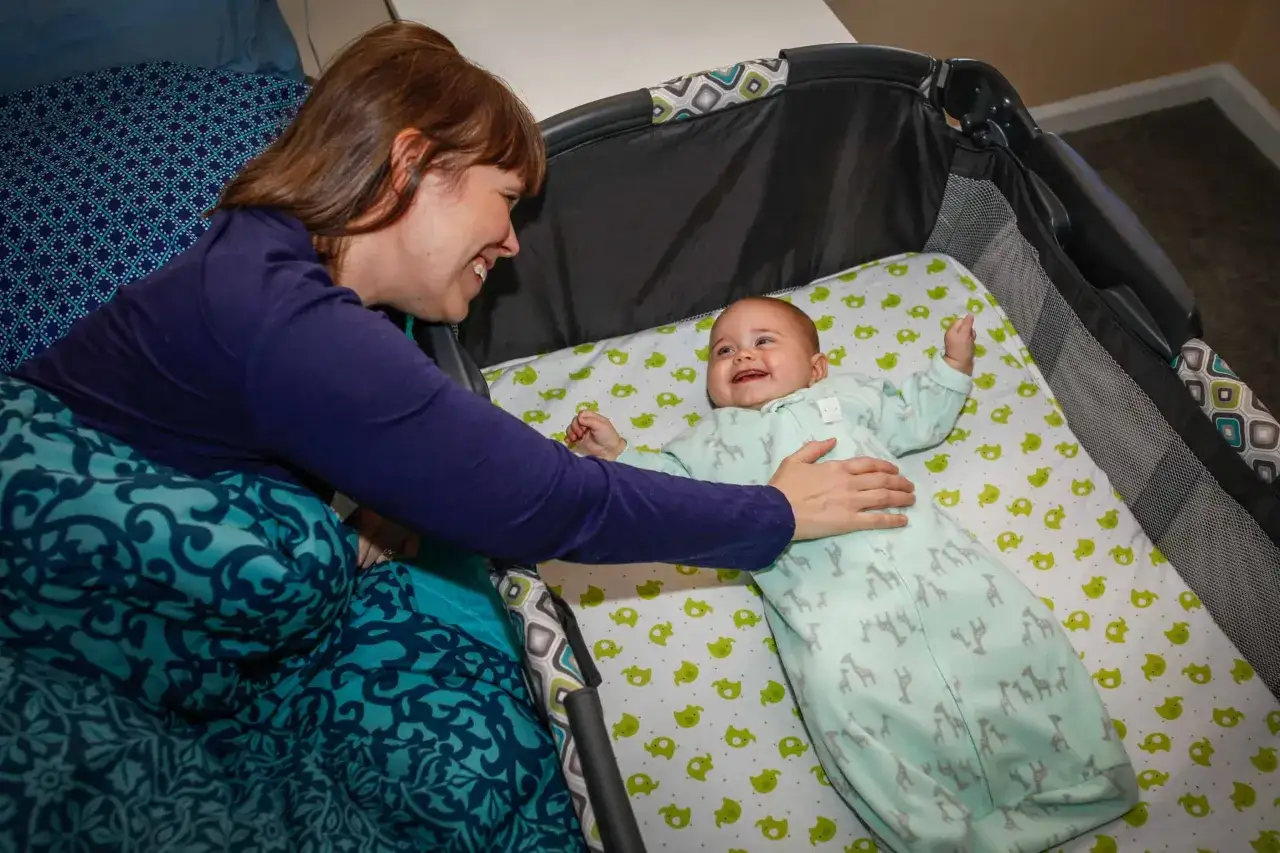 Madre sonr&iacute;e a su beb&eacute; que est&aacute; en la cuna, con un mameluco de jirafas. El beb&eacute; duerme de lado, feliz y con los brazos levantados.