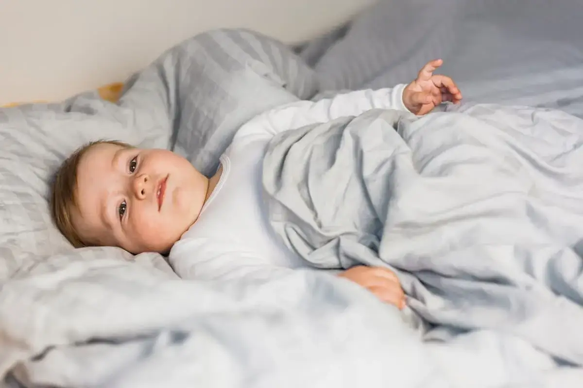 Un beb&eacute; sonriente yace en la cama, envuelto en mantas grises. &iquest;C&oacute;mo despertar a un beb&eacute; as&iacute;? Con ternura y paciencia.