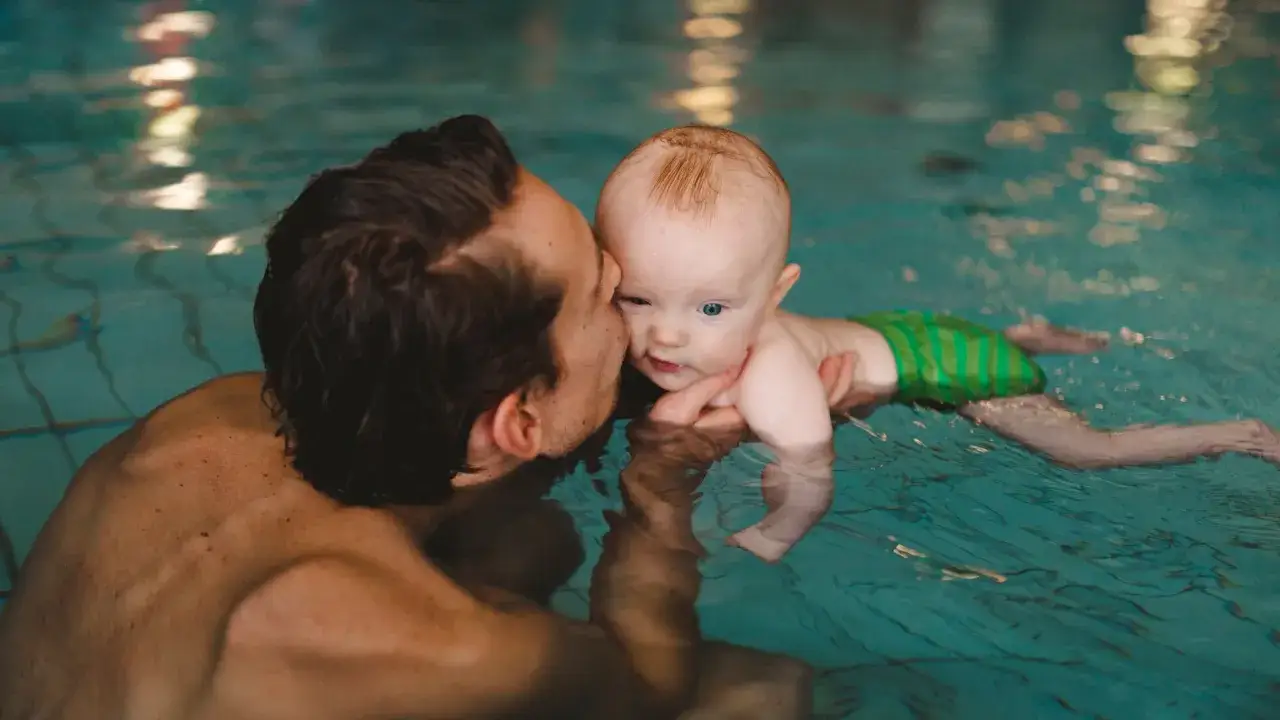 Padre besa a su beb&eacute; en la piscina, disfrutando de un momento tierno de nataci&oacute;n para beb&eacute;s.