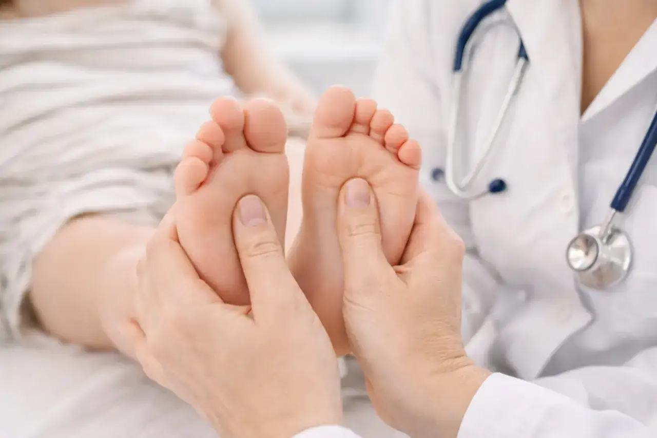 Manos de doctor examinando los pies de un ni&ntilde;o, evaluando posibles pies planos. El estetoscopio cuelga del cuello del m&eacute;dico.
