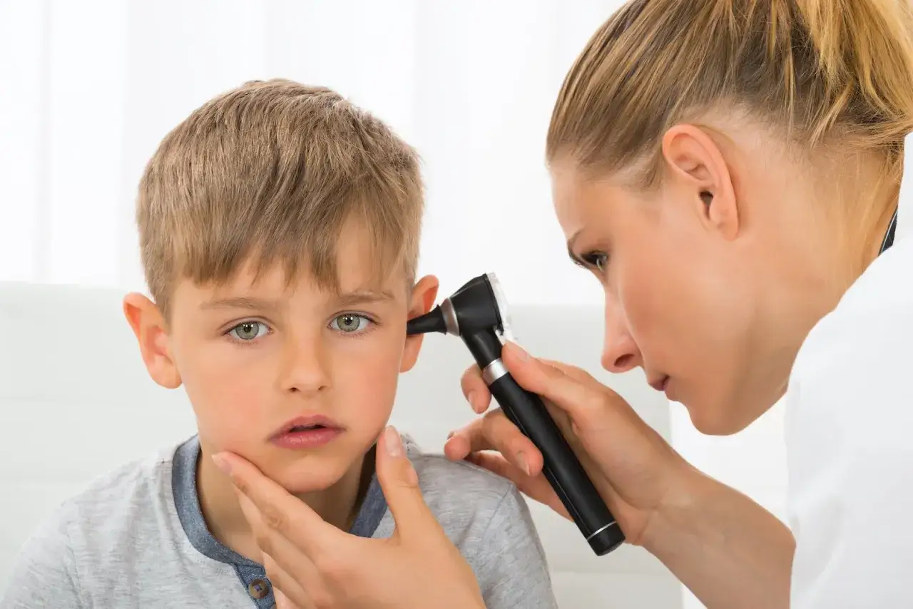 M&eacute;dico examina el o&iacute;do de un ni&ntilde;o con otoscopio. El ni&ntilde;o parece preocupado, quiz&aacute;s por la otitis.