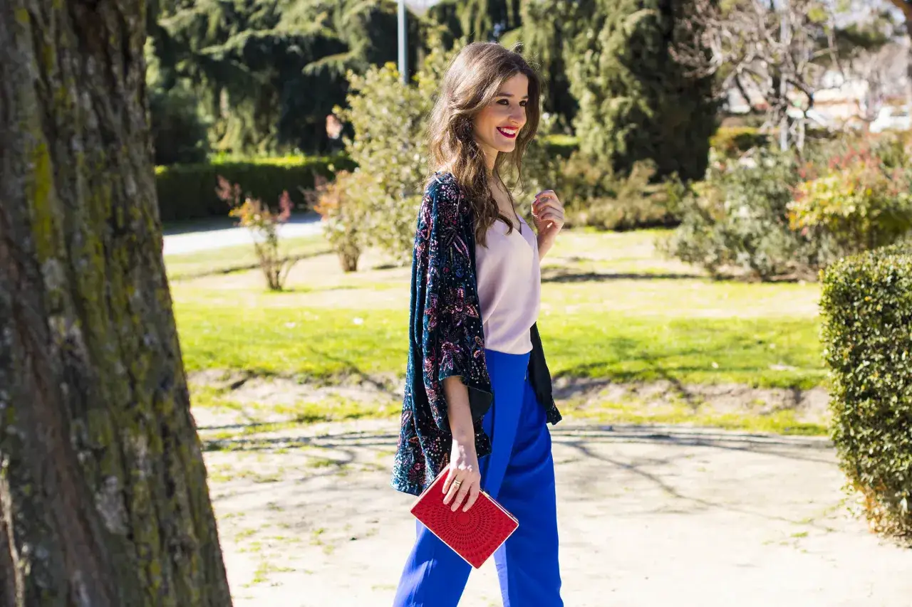 Una de las invitadas a la comuni&oacute;n, con un elegante kimono bordado, pantal&oacute;n azul y clutch rojo, pasea por un parque soleado.