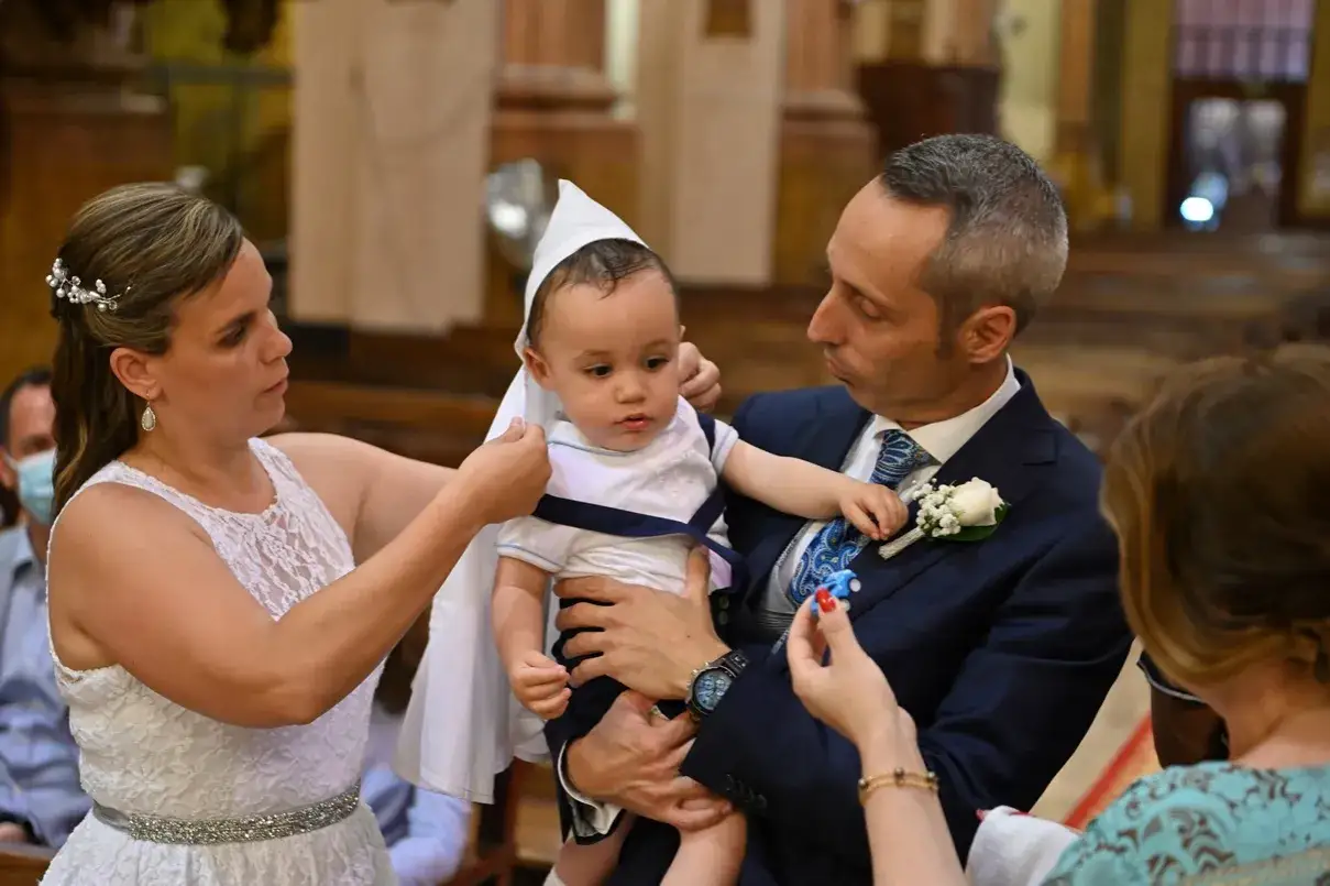 Padre y madre preparan al beb&eacute; para el bautizo en la iglesia. El padre sostiene al ni&ntilde;o, vestido con un traje blanco y azul, mientras la madre ajusta su gorro.
