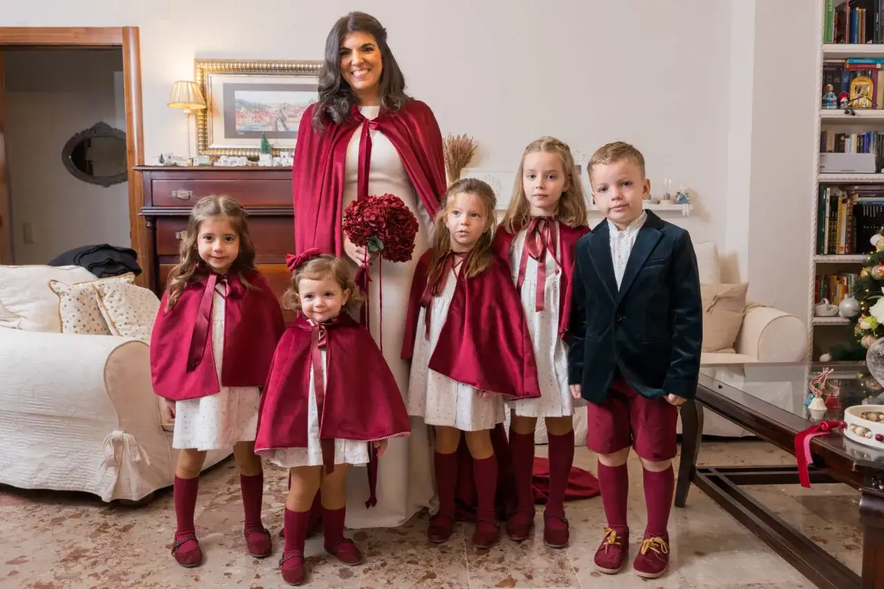 Novia sonriente con un ramo de flores rojas, rodeada de ni&ntilde;os con capas y trajes a juego, listos para una boda de invierno.