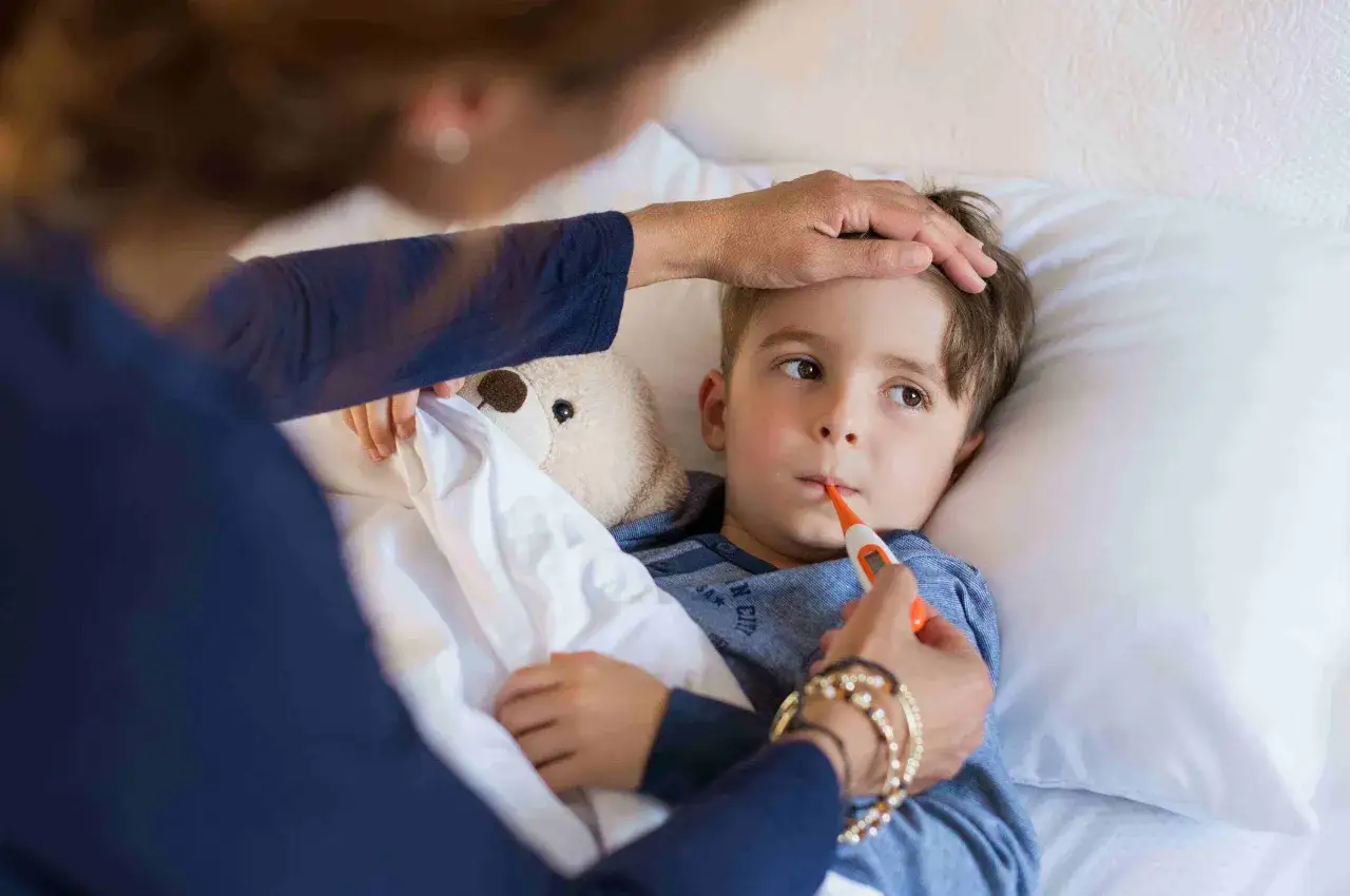 Ni&ntilde;o en cama con term&oacute;metro en la boca, su frente tocada por una mano. Posibles s&iacute;ntomas de neumon&iacute;a en ni&ntilde;os.