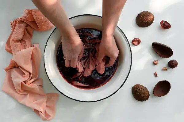 Manos sumergiendo tela en tinte oscuro para aprender como teñir ropa a mano. Cáscaras de aguacate alrededor.