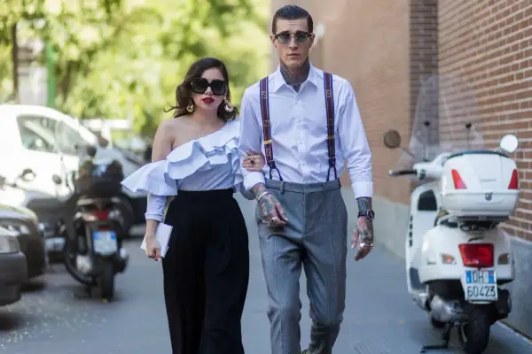 Pareja elegante camina, él es un invitado boda hombre sin traje, con tirantes y tatuajes visibles.