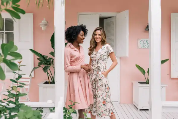 Dos mujeres posan frente a una casa rosa. Una lleva un vestido rosa liso, la otra uno floral. Ideas para como ir a una boda informal mujer.
