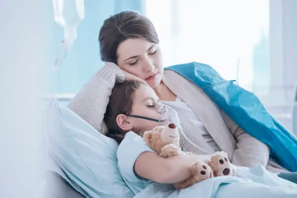 Niña con mascarilla de oxígeno y oso de peluche, su madre la abraza. Posibles síntomas de neumonía en niños.