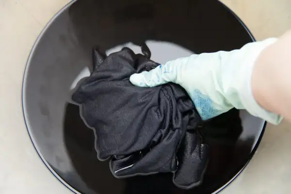 Mano con guante sumerge tela negra en líquido oscuro para recuperar color de ropa oscura.