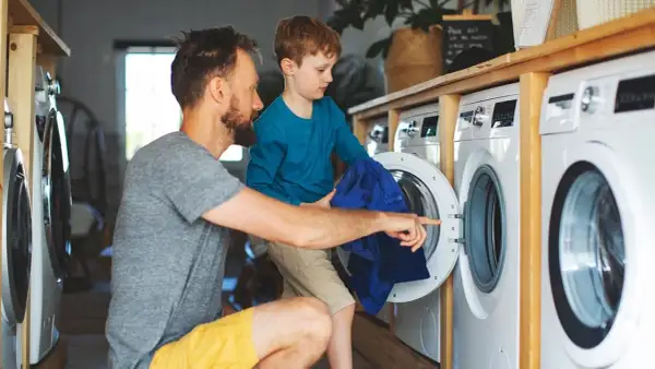 Padre e hijo aprenden como lavar ropa de deporte. El niño ayuda a meter la ropa azul en la lavadora.