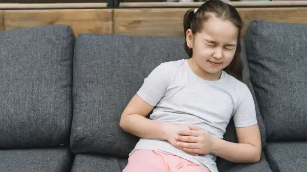 Niña con dolor de barriga en niños cuando preocuparse. Se agarra el abdomen con expresión de malestar sentada en un sofá.