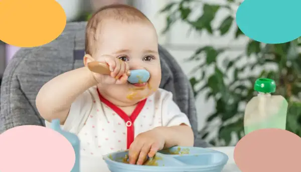 Un bebé de 12 meses, con la cara manchada de comida, intenta alimentarse solo con una cuchara azul.