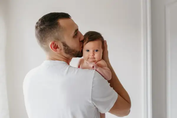 Un padre besa la cabeza de su bebé, que mira a la cámara con sus grandes ojos azules.